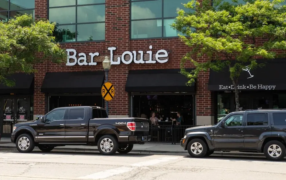 Bar Louie exterior in daylight: white 'Bar Louie' lettering on a brick building with black awnings, two black SUVs/pickups parked in front.